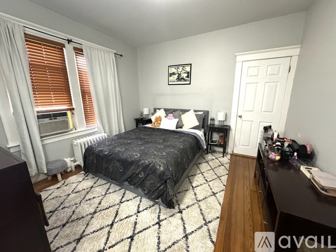 A bedroom with a black and white checkered rug, a bed with a black comforter, a desk with a computer, and a window with blinds.