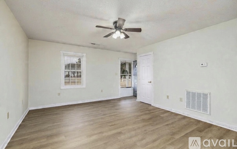 A spacious room with a ceiling fan and light, hardwood floors, and a white door leading to another room.