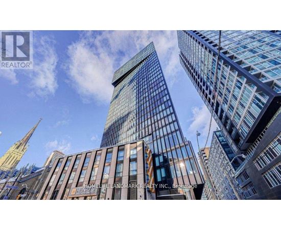 A tall building with a glass facade stands in front of a skyline of other buildings.