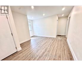 A room with wooden flooring and white walls, with a door on the left and a window on the right.