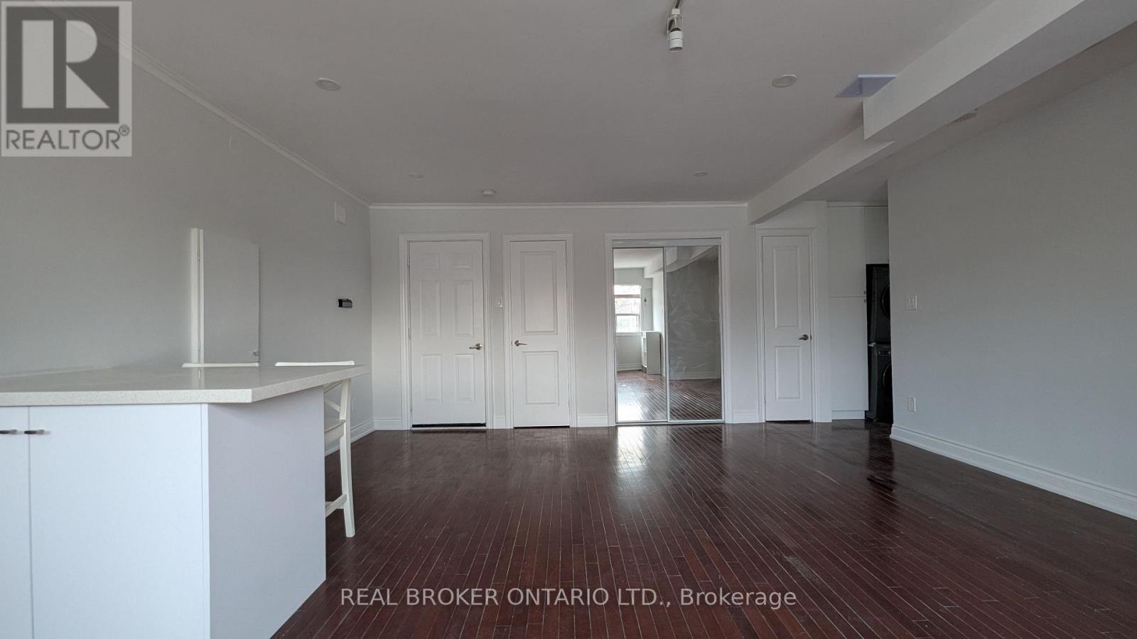A spacious room with wooden floors and white walls, featuring a kitchen area with white cabinets.
