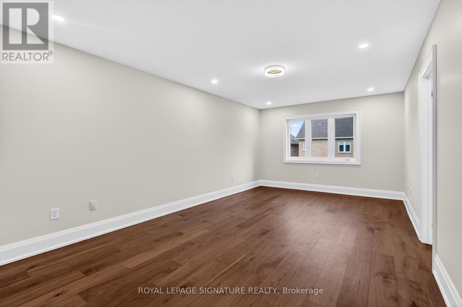 A spacious room with wooden flooring and a window showing a view of a house.