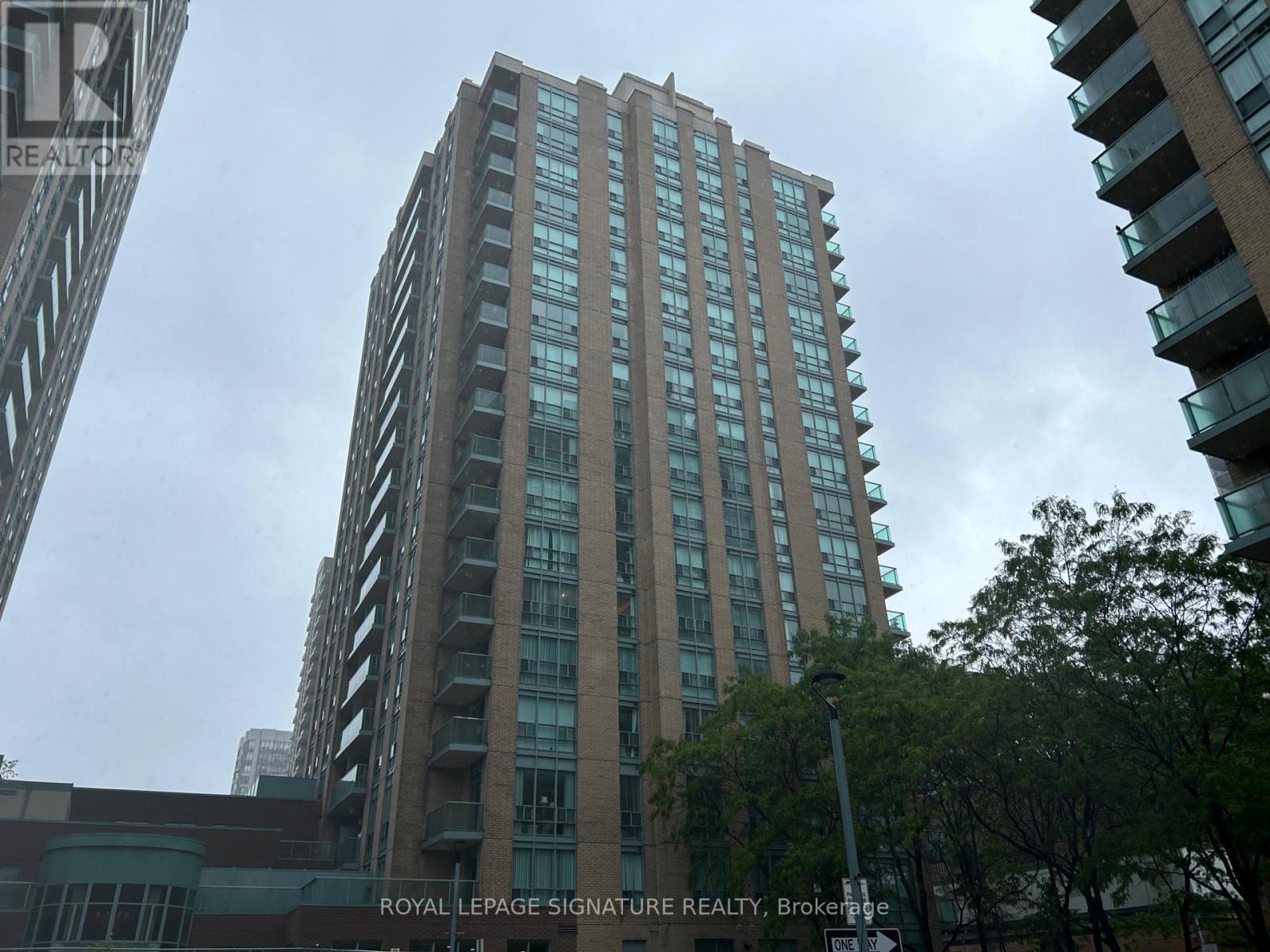 A tall building with many windows is in the foreground with trees and another building in the background.