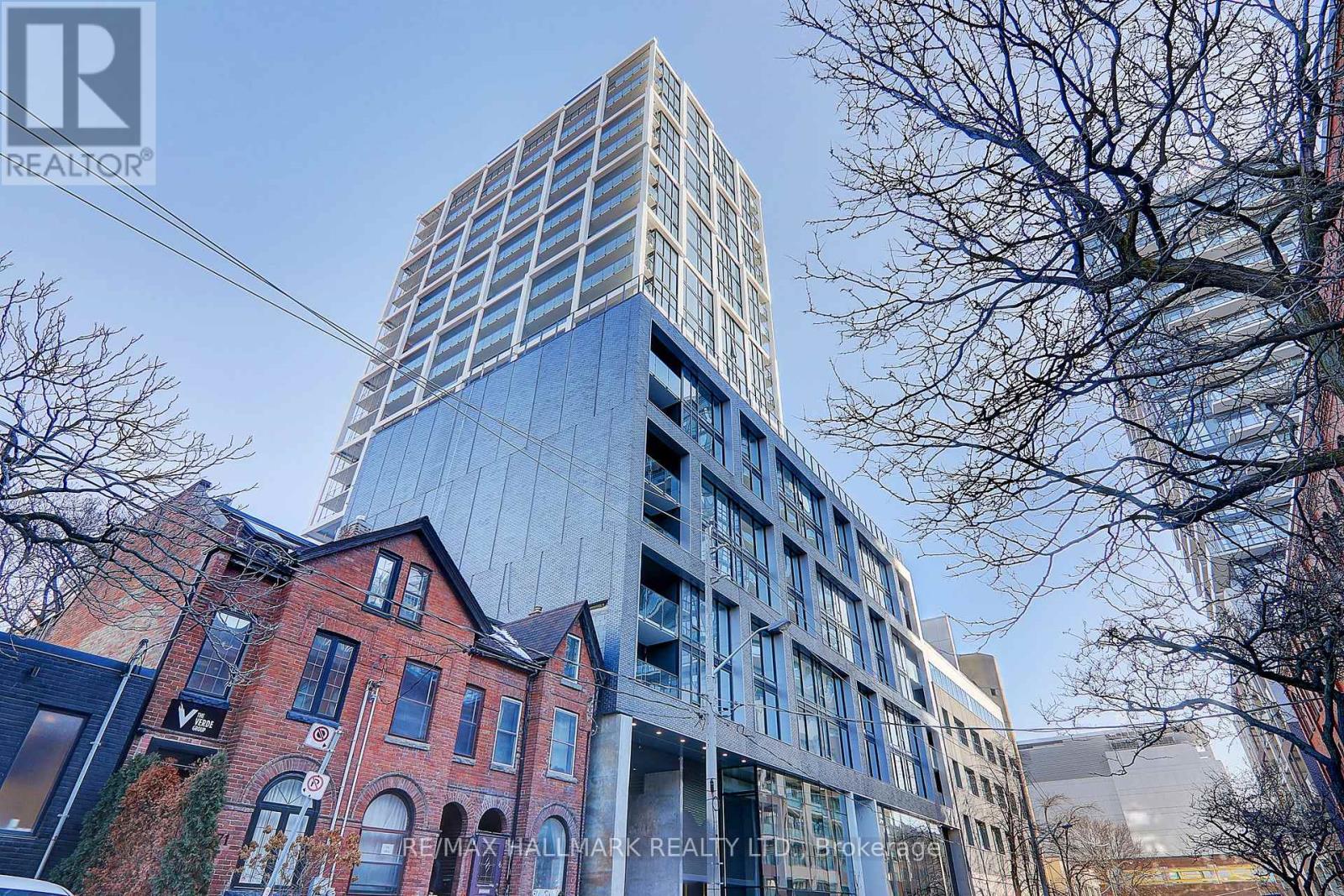 A tall glass building stands behind a red brick building with trees in the foreground.