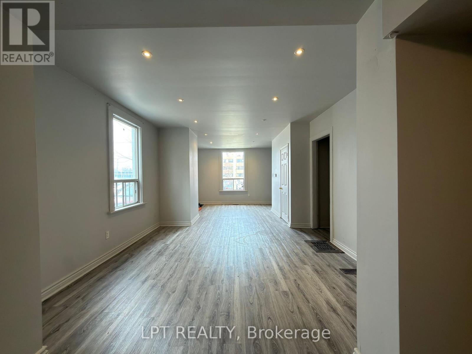 A spacious room with wooden flooring and white walls, with a window on the left side.