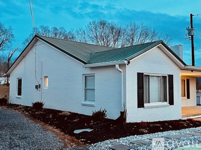 A small white house with a green roof and a gravel driveway.