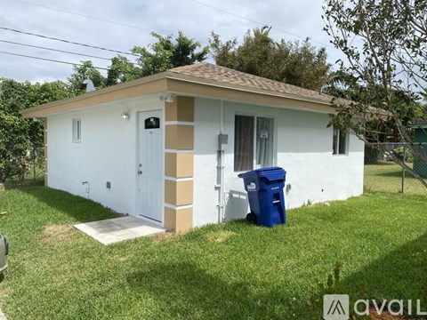 A small white house with a brown roof and a blue trash bin in front.