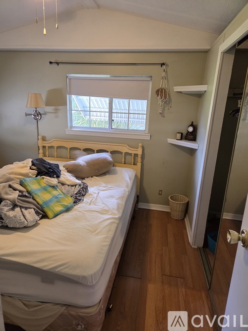 A bedroom with a bed, a window with blinds, a nightstand with a lamp, and a shelf with a clock and a plant.
