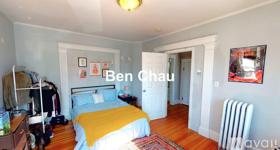 A bedroom with a bed, a chair, a nightstand, a dresser, a window, a wall-mounted shelf, a rug, a painting, a lamp, a door, a wall-mounted heater, and