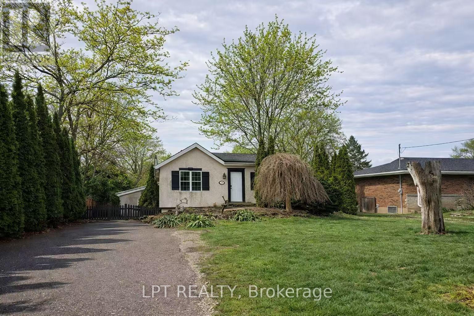 A small white house with a brown roof is surrounded by greenery and a gravel driveway.