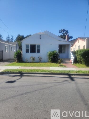 A white house with a brown roof is situated on a street with a sidewalk.