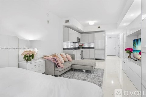 A modern bedroom with a large bed and a kitchen area in the background.