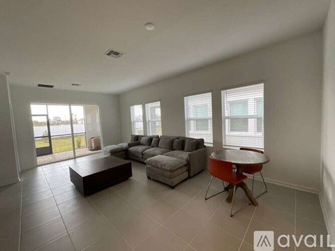 A spacious living room with a grey sofa set, a red coffee table, and a large window with blinds.