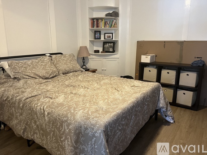 A bed with a grey bedspread and pillows is in the foreground with a bookshelf and a lamp behind it.