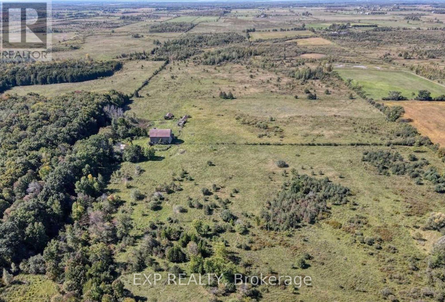 An aerial view of a large, open field with a small house in the distance and a forested area to the left.