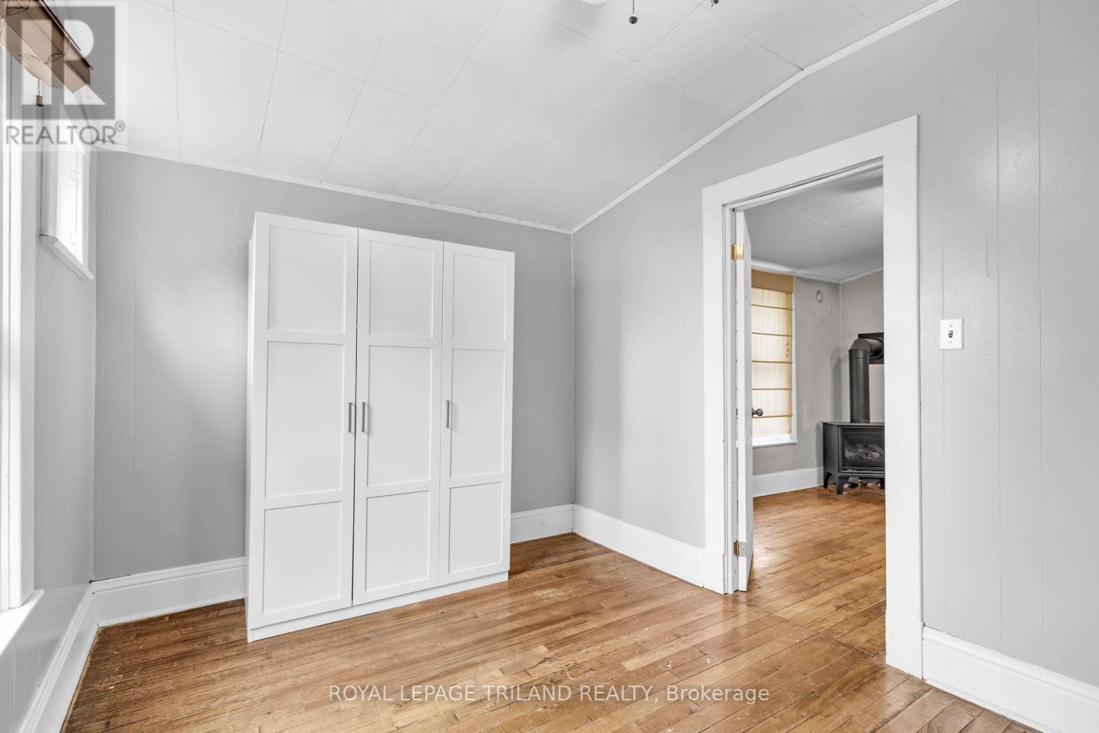 A room with white walls and wooden floors, with a white wardrobe and a doorway leading to another room.