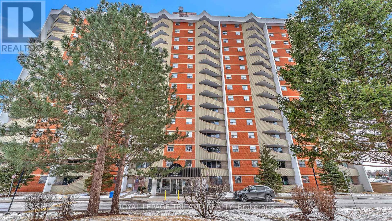A tall apartment building with a red and orange facade is surrounded by trees and a car is parked in front.