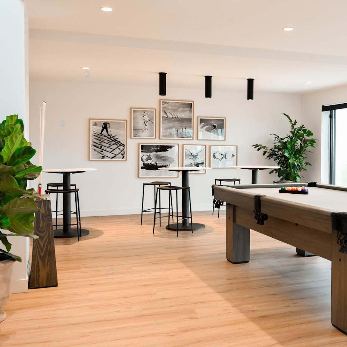 A pool table is in the middle of a room with a light wood floor and a white wall with pictures hanging on it.