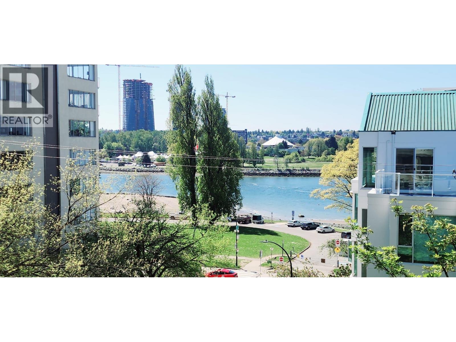 A realtor sign is in the foreground of a cityscape with a lake and greenery in the background.