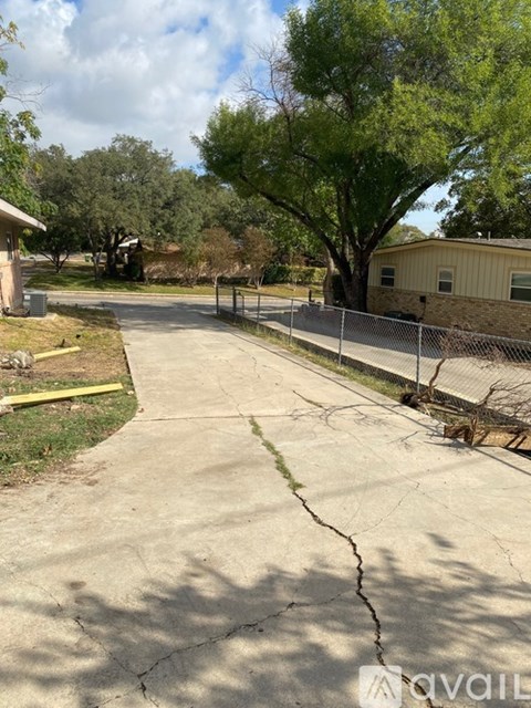 A cracked driveway leads to a house with a fence and trees in the background.