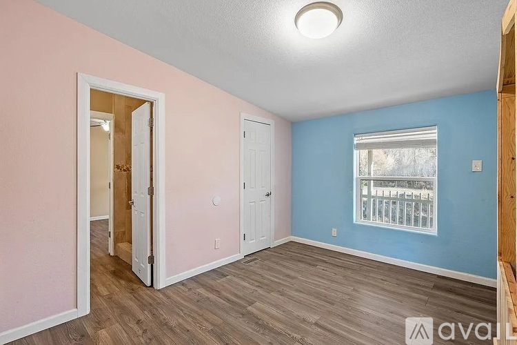 A room with a doorway leading to another room, a window with a view of trees, and a wooden floor.