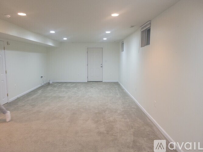 A spacious room with beige carpeting and white walls, with a door on the right and a small window on the left wall.