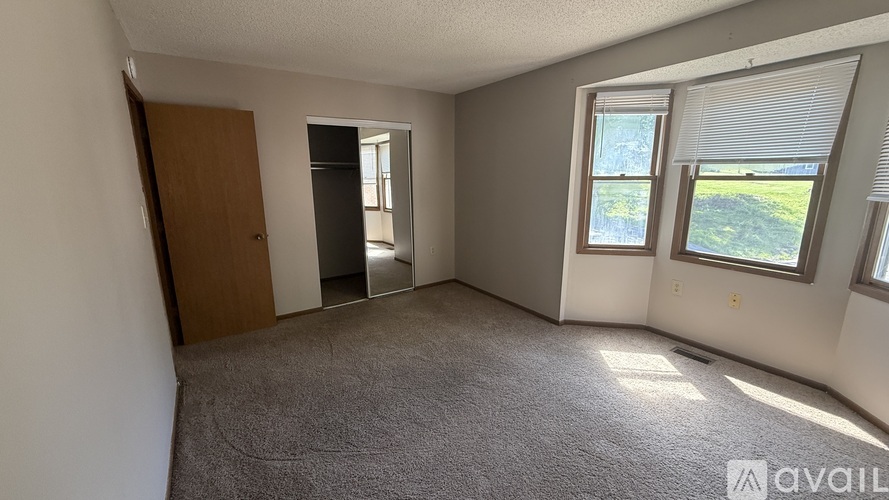 A room with a carpeted floor, a wooden door, a window with blinds, and a wall with a reflection of a building.