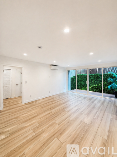 A spacious room with wooden flooring and a large window overlooking a green area.