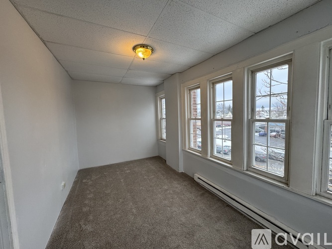 A room with a carpeted floor, a window with a view of a parking lot, and a ceiling light.