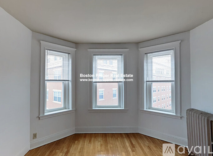 A room with wooden floors and three windows, one of which has a view of a building.