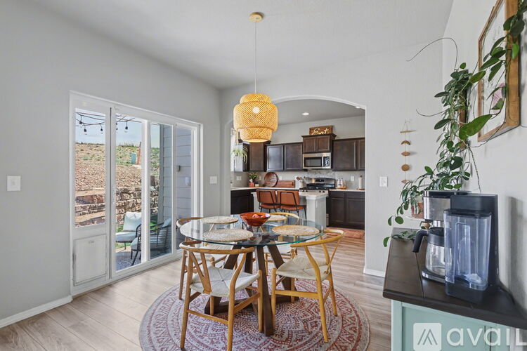 A dining room with a glass table and chairs, a hanging light fixture, and a kitchen area in the background.