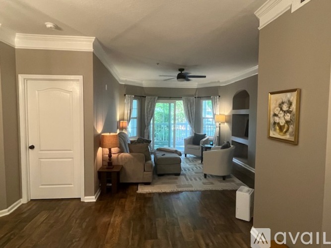 A living room with a white door, a couch, a chair, a table, a ceiling fan, and a painting on the wall.