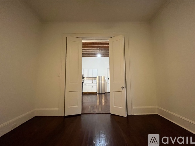 A room with white walls and a wooden floor leading to a kitchen.