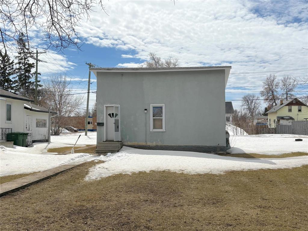 A small white building with a door and windows sits in a grassy area with snow on the ground.