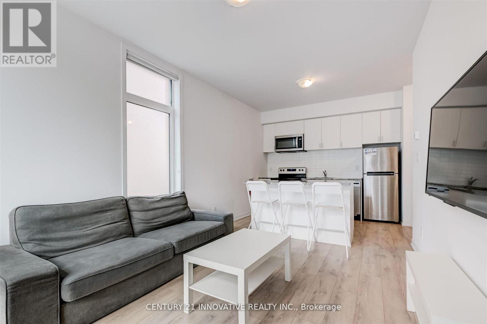 A grey couch sits in a white living room with a kitchen area in the background.