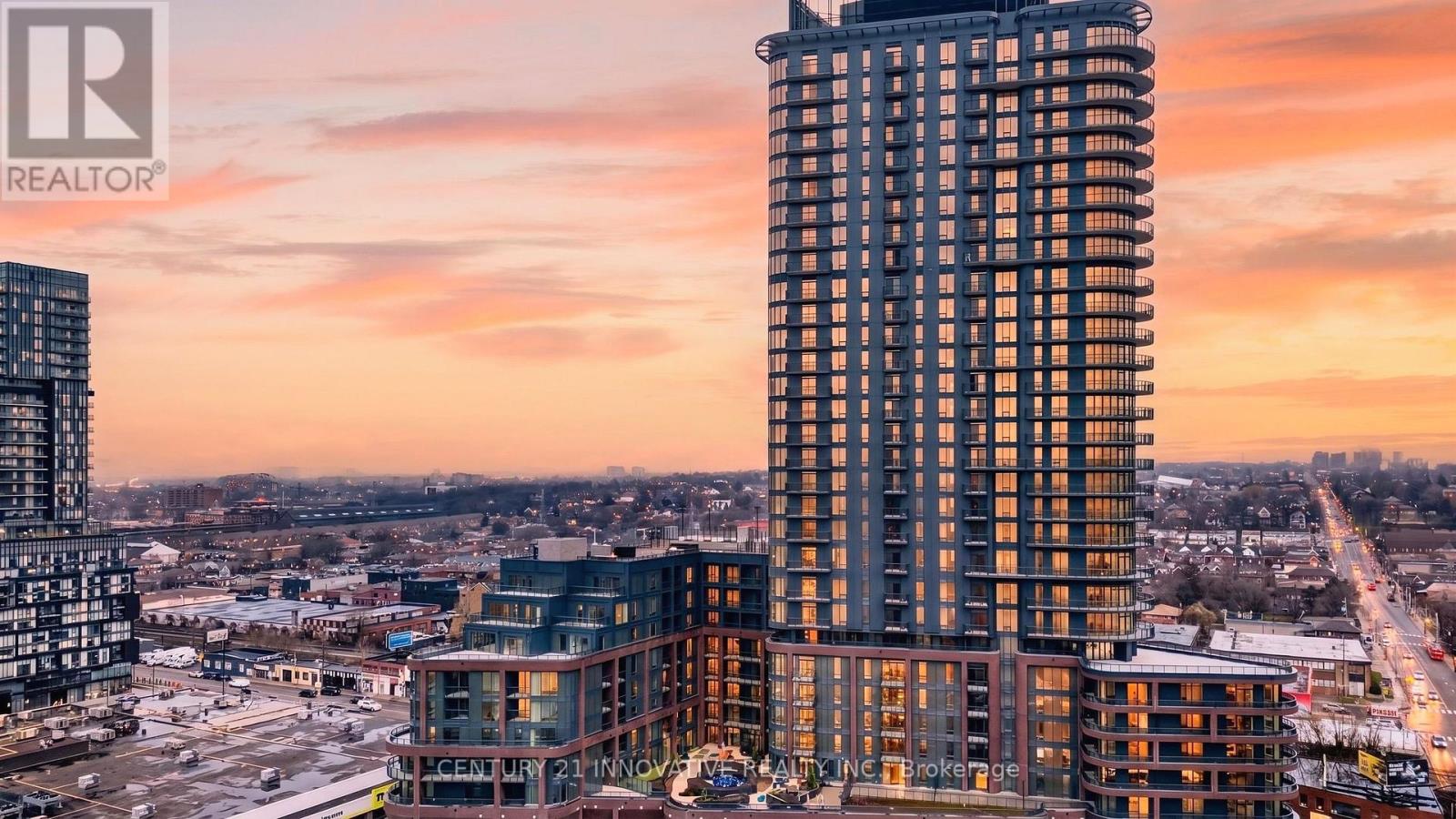 A tall building with a glass facade stands in the foreground of a cityscape at sunset.