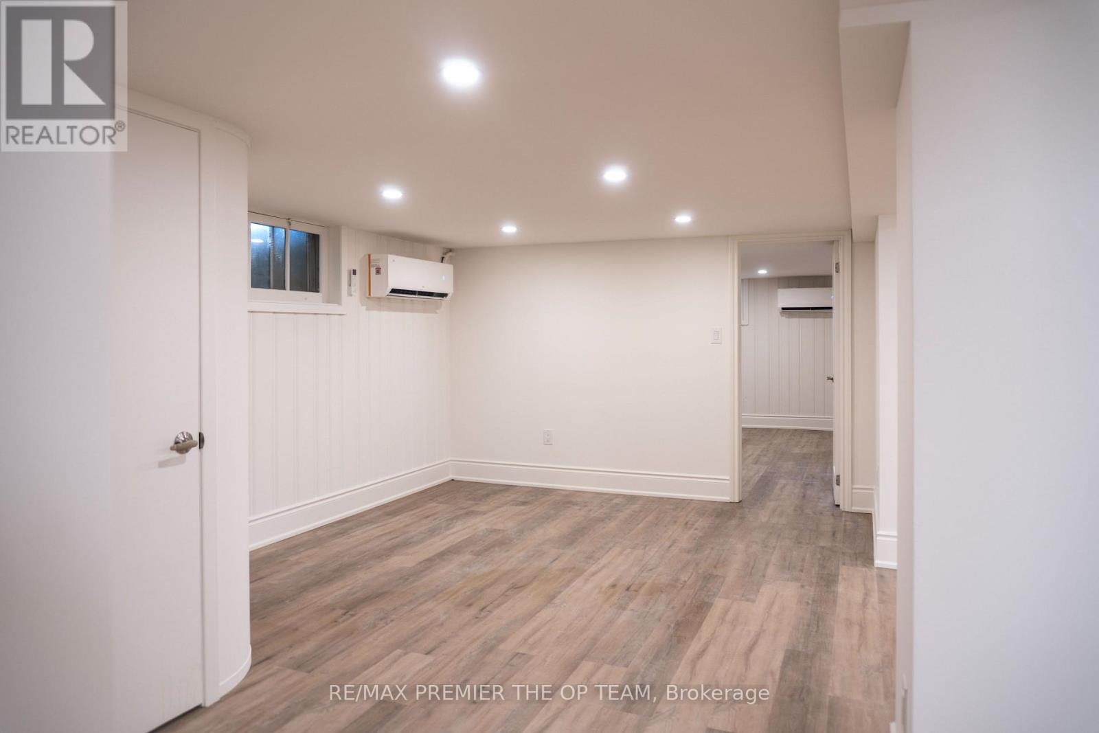 A spacious room with wooden flooring and white walls, featuring a door on the left and a window on the right.
