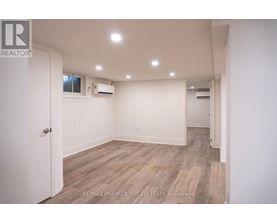 A spacious room with wooden flooring and white walls, featuring a door on the left and a window on the right.