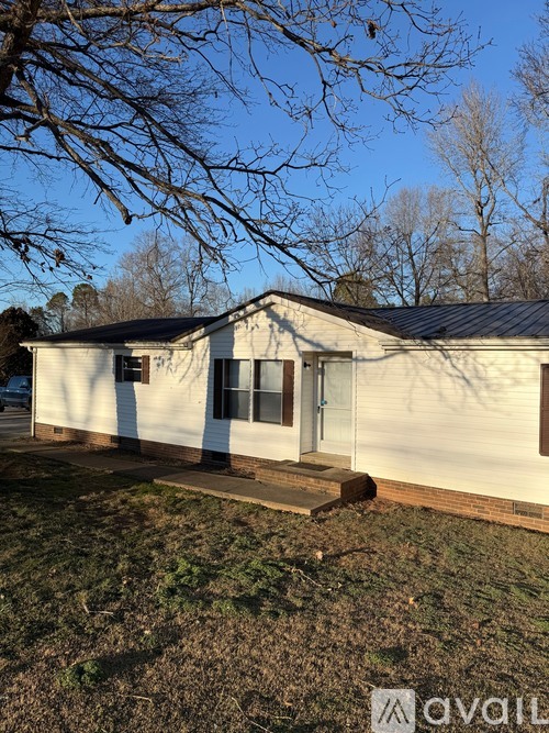 A house with a white siding and a black roof is surrounded by a grassy area and leafless trees.