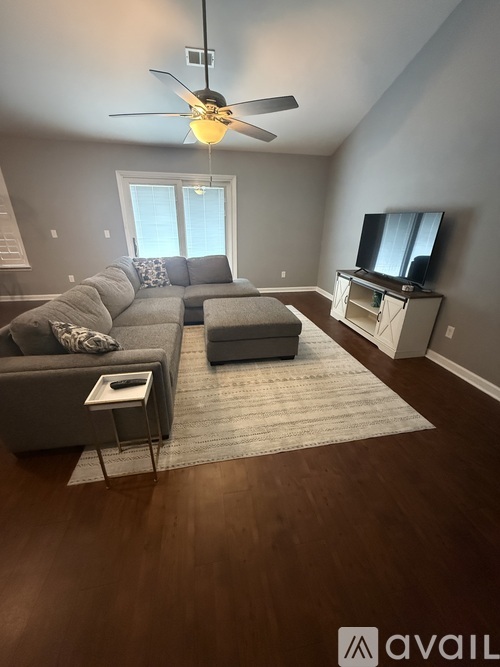 A living room with a grey couch, a white coffee table, a grey ottoman, a television, and a fan.