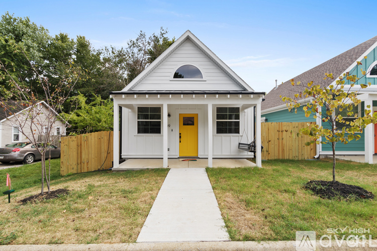 A small white house with a yellow door is surrounded by a wooden fence and has a small tree in front of it.