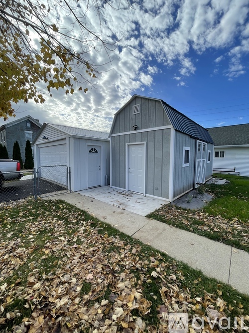 A grey barn-like building with a white door is surrounded by a fence and a pile of leaves.