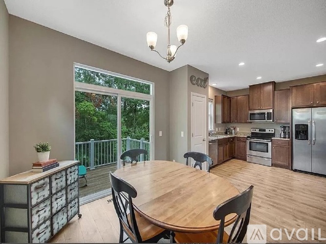 A dining room with a wooden table and chairs, a kitchen in the background, and a balcony with a view of trees.