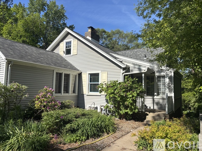 A house with a grey siding and a white window is surrounded by greenery.