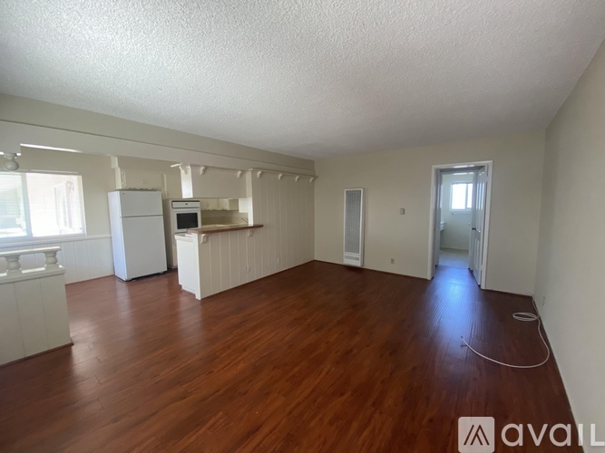 A room with wooden floors and white walls, with a refrigerator and a window.