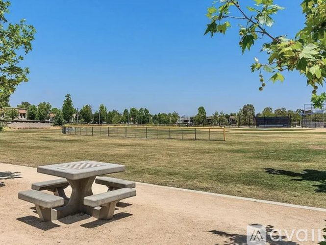 A picnic table is in the foreground of a park with a fence and trees in the background.
