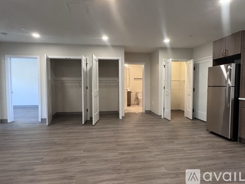 A spacious room with wooden flooring and white walls, featuring a refrigerator on the right and several open doors.