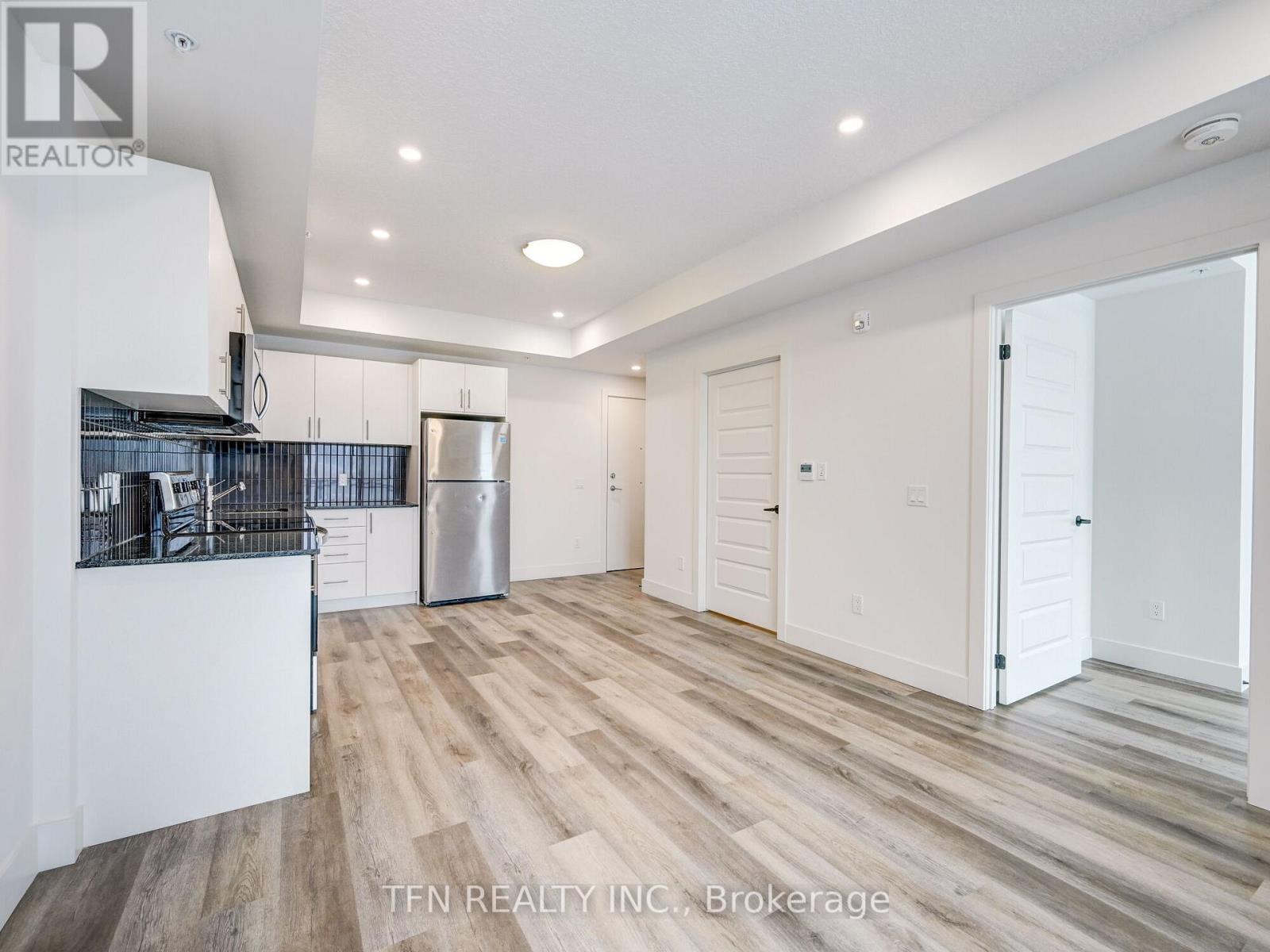 A spacious room with wooden flooring and white walls, featuring a kitchen area with a refrigerator and cabinets.