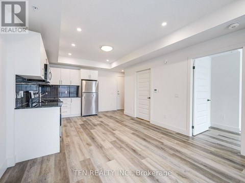 A spacious room with wooden flooring and white walls, featuring a kitchen area with a refrigerator and cabinets.