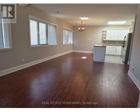A large empty room with wood flooring and a kitchen in the background.